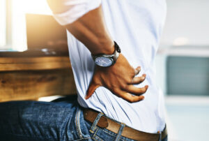 Image of an office with a businessman standing, hands on waist, experiencing lower back pain on the left side.